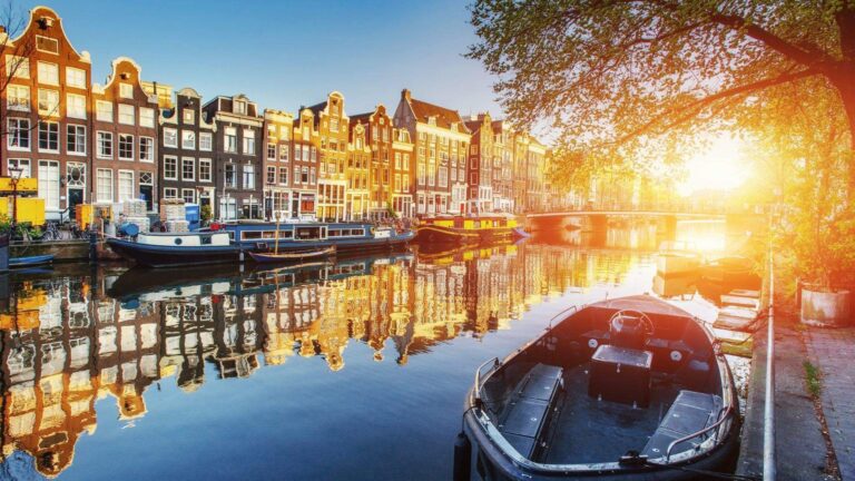 Saulėlydžio apšviestas Amsterdamo kanalas su istoriniais namais ir priekyje stovinčia valtimi