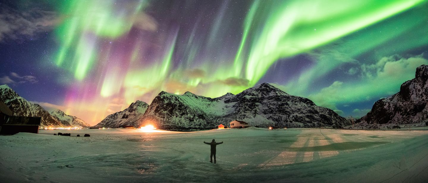 Žmogus išskėstomis rankomis stovi snieguotame lauke po ryškia šiaurės pašvaiste Norvegijos kalnuose