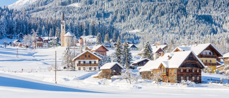 Snieguotas Austrijos kaimas su mediniais namais ir bažnyčia apsnigtų miškų apsuptyje