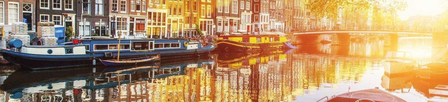 Saulėlydžio apšviestas Amsterdamo kanalas su istoriniais namais ir priekyje stovinčia valtimi