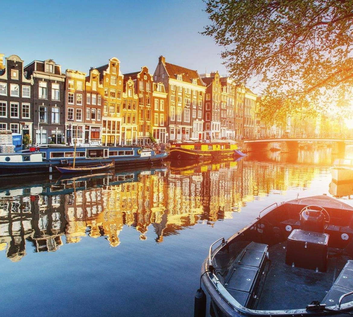 Saulėlydžio apšviestas Amsterdamo kanalas su istoriniais namais ir priekyje stovinčia valtimi