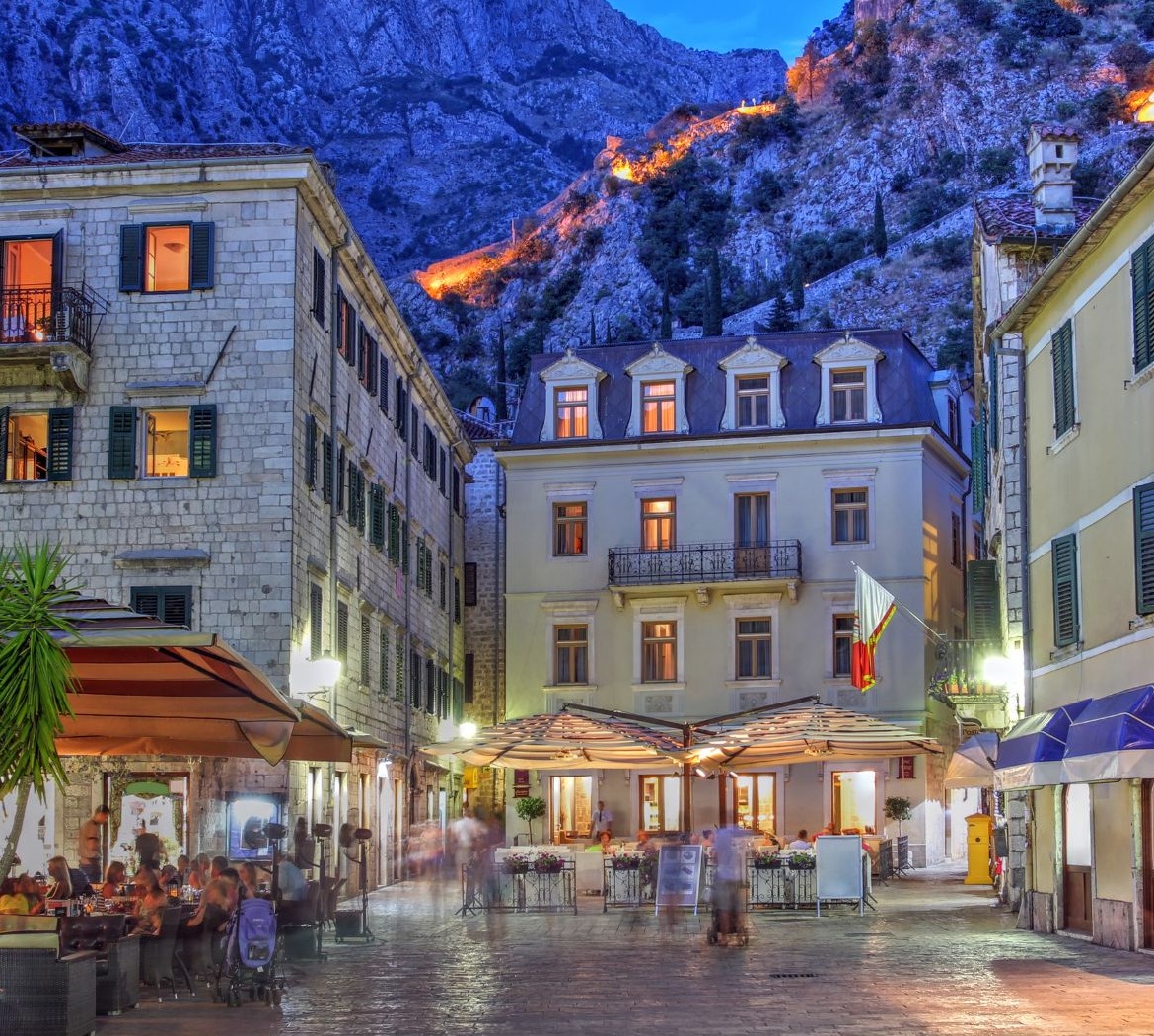 Vakarinis Kotoro senamiestis su akmeniniais pastatais, lauko kavinėmis ir apšviesta tvirtovės siena